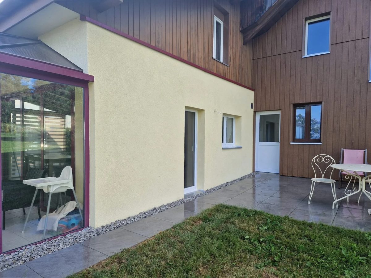 Façade de maison avec un crépi neuf de teinte ivoire, entre bardage en bois et véranda